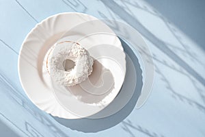 Donut covered with white glaze and sprinkled with coconut flakes on a white plate on blue background under natural light