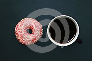 Donut in pink glaze and cup of coffee on dark background, view from above