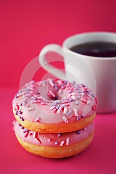 Donuts in pink glaze with sprinkles next to a white cup of coffee