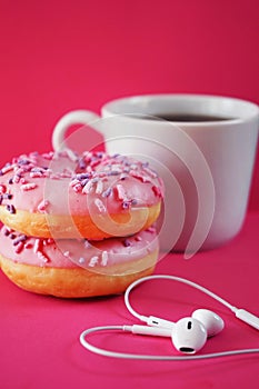 Donuts in pink glaze with sprinkles next to a white cup of coffee and headphones laid out in the shape of a heart