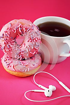 Donuts in pink glaze with sprinkles next to a white cup of coffee and headphones laid out in the shape of a heart