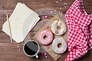 Donuts with sprinkles hearts, cup of coffee and old paper sheet, wooden background, top view