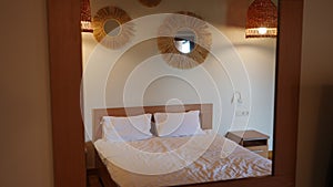 Double bed in hotel room with white clean bedding reflecting in mirror on wall at tourist resort. Comfortable furniture