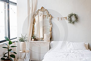 A double bed with white linens and pillows in the bedroom decorated for Christmas and New Year. Christmas wreath near the mirror