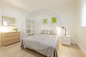 Double bedroom with white wooden headboard, gray bedspread and cushions, white bedside table with folding lamp and oak chest of