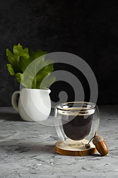 Double bottom glass cup with coffee on wooden stand with acorn and white milk jug with green oak leaves in low key