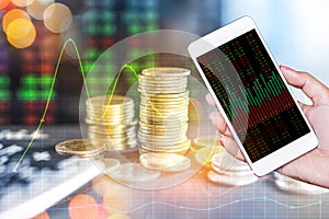 Double exposure hand holding smart phone and stack of coins over stock market screen