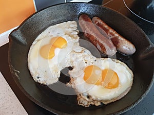 Double Fried Eggs Sunny Side Up, fried sausages on a cast iron frying pan on a cooktop background