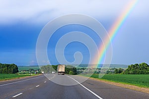 Double rainbow in the blue cloudy dramatic sky over green field and a road illuminated by the sun in the country side