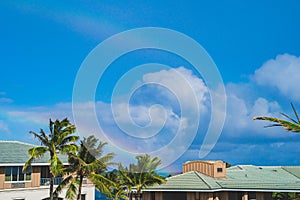 Double rainbow in blue sky above palm trees and rooftops in tropical paradise