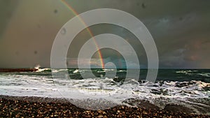 Double rainbow in the sky with clouds after the storm, in the foreground the sea waves hit the pebble beach, behind you