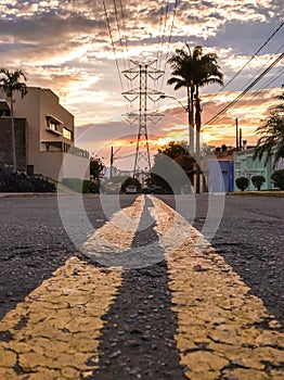 Double yellow lines on the street leading to a power line tower against sunset sky