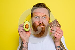 Doubter man with beard and tattoos holds avocado and chocolate