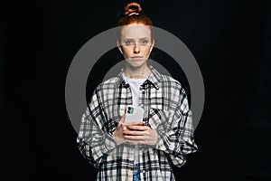 Doubtful sad young woman wearing wireless earphones typing online message on black background