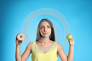 Doubtful woman choosing between apple and doughnut on light blue background