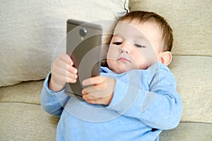 Doubting toddler baby boy is sitting with a phone on the sofa in the livi