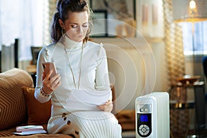 Doubting woman near radiator with smartphone and utility bill