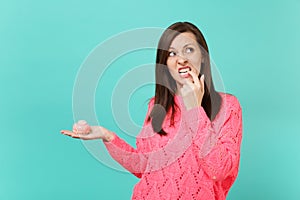 Doubting young woman in knitted pink sweater looking up, holding in hand cake, gnawing nails isolated on blue turquoise