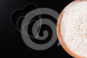 Dough cutter in shape of heart and a bowl with flour on black table. Valentines Day baking background