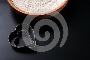 Dough cutter in shape of heart and a bowl with flour on black table. Valentines Day baking background