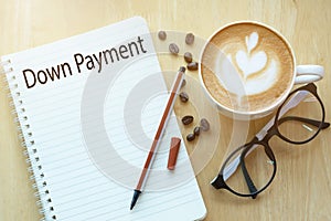 Down payment word on notebook with glasses, pencil and coffee cup on wooden table. Business concept