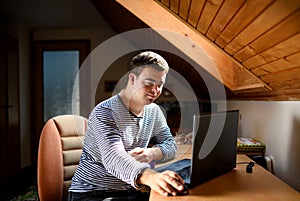 Down syndrome adult man sitting indoors in bedroom at home, using laptop.