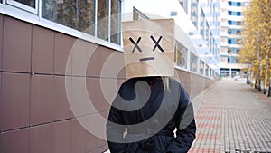 Downcast scared paper bag face walking along street. Woman wearing bread bag with x cross eyes