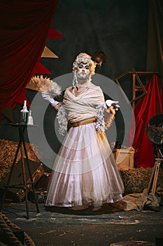 Drag queen, man in female white dress and bright makeup over dark retro circus backstage background.