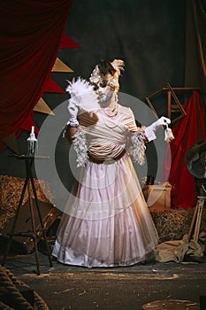 Drag queen, man in female white dress and bright makeup over dark retro circus backstage background.