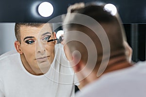 Drag queen performer looking in the mirror while applying mascara on his eyelashes.