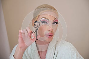 Drag queen person curling the lashes with a eyelash curler and wearing bathrobe.