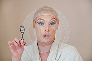 Drag queen person with a eyelash curler and wearing bathrobe looking at camera.