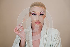Drag queen person with lip gloss brush wearing bathrobe and looking at camera.