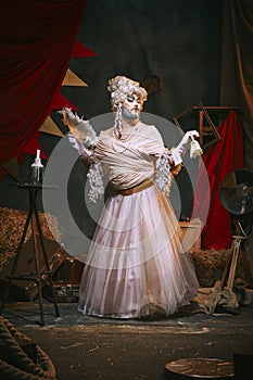Drag queen in white dress and bright makeup over dark retro circus backstage background. Man in female appearance