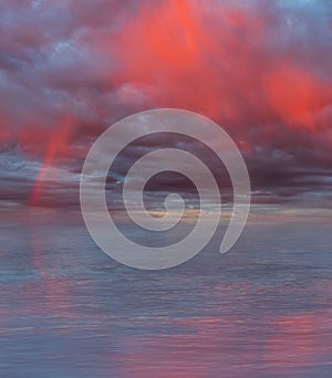 Dramatic cloudy sky with rainbow and red rain curtains reflecting on sea