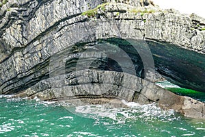 Dramatic coastal cliff rock layers curve, forming cave in teal aqua ocean waves on Loop Head Peninsula, County Clare, Ireland.