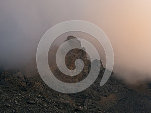 Dramatic fog among giant rocky mountains. Ghostly atmospheric view to big cliff in cloudy sky. Low clouds and beautiful rockies.