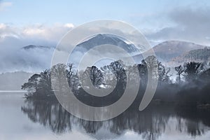 Dramatic landscape image looking across Derwentwater in Lake District towards Catbells snowcapped mountain with thick fog rolling