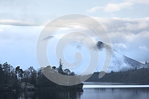 Dramatic landscape image looking across Derwentwater in Lake District towards Catbells snowcapped mountain with thick fog rolling