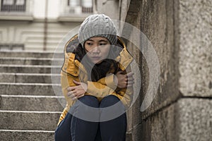 Dramatic lifestyle portrait of young attractive sad and depressed Chinese woman in winter hat sitting outdoors on street corner