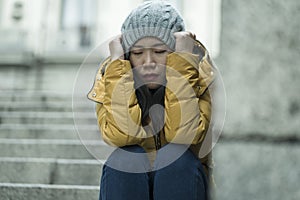 Dramatic lifestyle portrait of young attractive sad and depressed Chinese woman in winter hat sitting outdoors on street corner