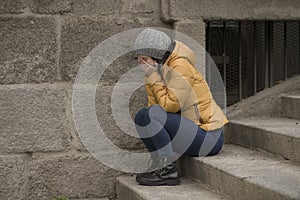 Dramatic lifestyle portrait of young attractive sad and depressed Chinese woman in winter hat sitting outdoors on street corner