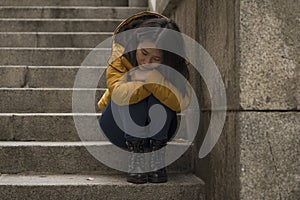 Dramatic lifestyle portrait of young attractive sad and depressed Japanese woman in winter jacket sitting outdoors on street