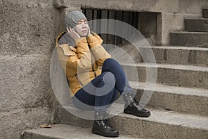 Dramatic lifestyle portrait of young attractive sad and depressed Korean woman in winter hat sitting outdoors on street corner