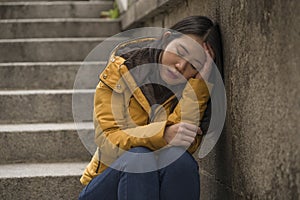 Dramatic lifestyle portrait of young attractive sad and depressed Korean woman in winter jacket sitting outdoors on street corner