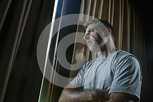 Dramatic light portrait of young sad and depressed attractive man at home looking through room window thoughtful and pensive lost