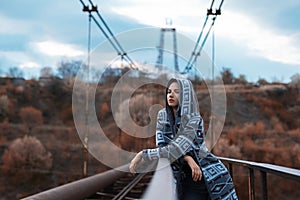 Dramatic outdoor portrait of young teenage hooded girl standing on abandoned old rusty bridge with perspective background.