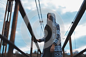 Dramatic outdoor portrait of young teenage hooded girl standing on abandoned old rusty bridge with perspective background.