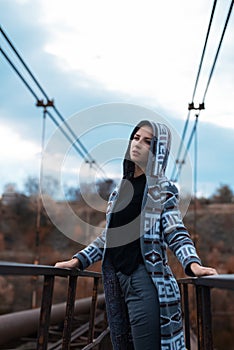 Dramatic outdoor portrait of young teenage hooded girl standing on abandoned old rusty bridge with perspective background.