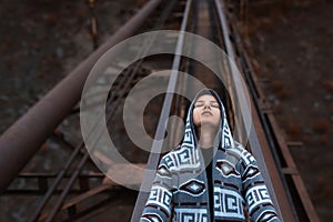 Dramatic outdoor portrait of young, thoughtful hooded girl, lying on old rusty bridge. Perspective concept.